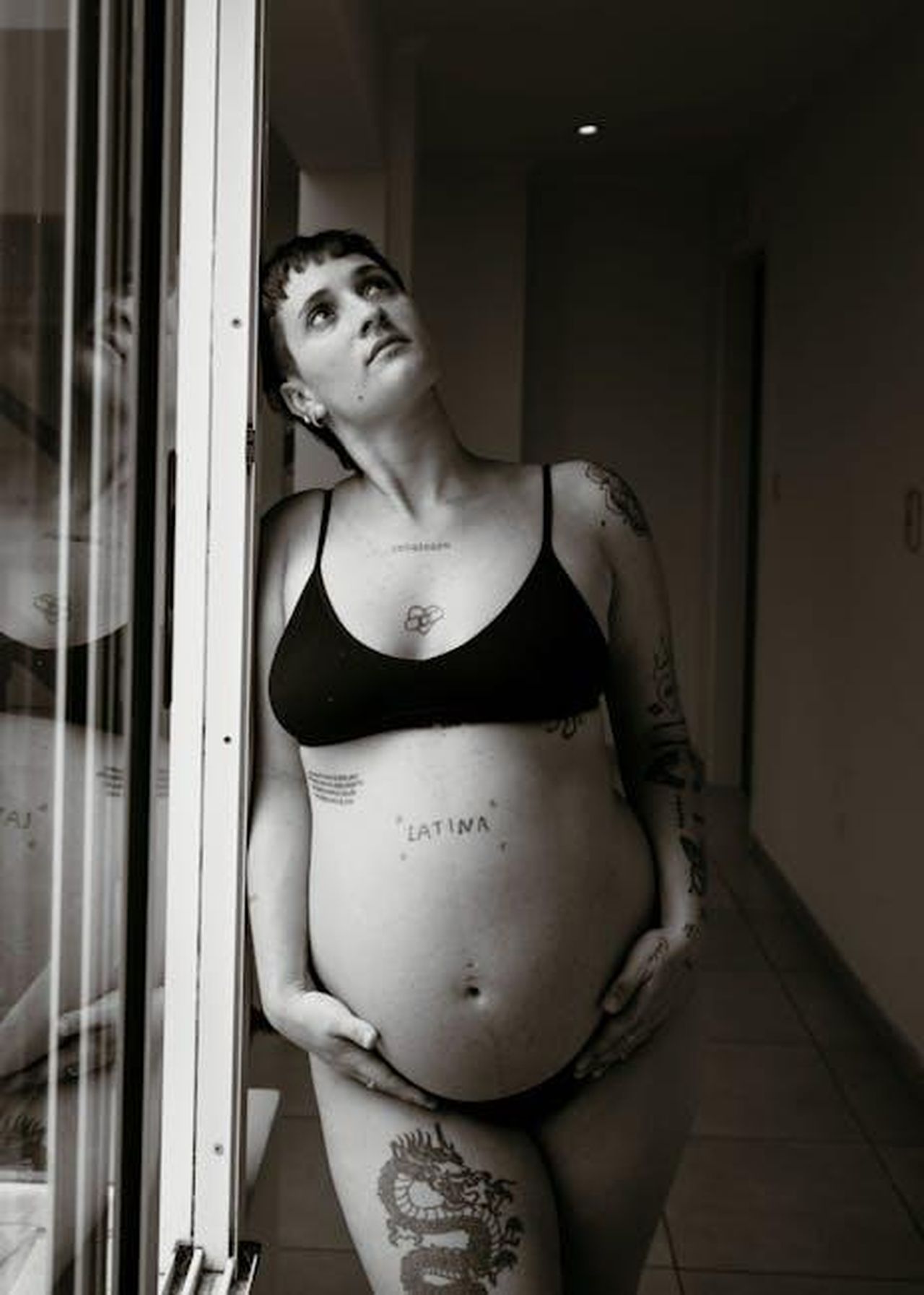 Riflessioni_sulla_bellezza_e_lautocritica_Yss3RevYg0 - Confidenze24 Source: Natalia Olivera - pexels - A pregnant woman with tattoos gazes thoughtfully, wearing a black bra indoors.