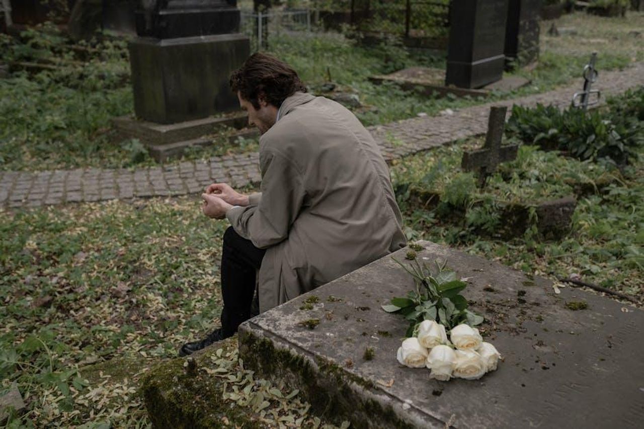 Riflessioni_sulla_vita_e_la_morte_PLGiqmld7q - Confidenze24 Source: cottonbro studio - pexels - A man in a trench coat sits grieving at a cemetery, white roses on a tombstone.