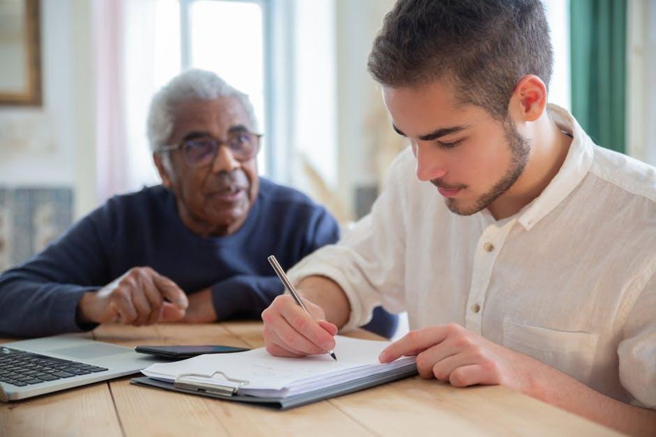 Rimani_Connesso_per_Alleviare_lo_Stress_mJ8cynWtWB - Confidenze24 Source: Kampus Production - pexels - A young man aids a senior citizen with writing at a wooden table indoors, fostering social assistance and connection.