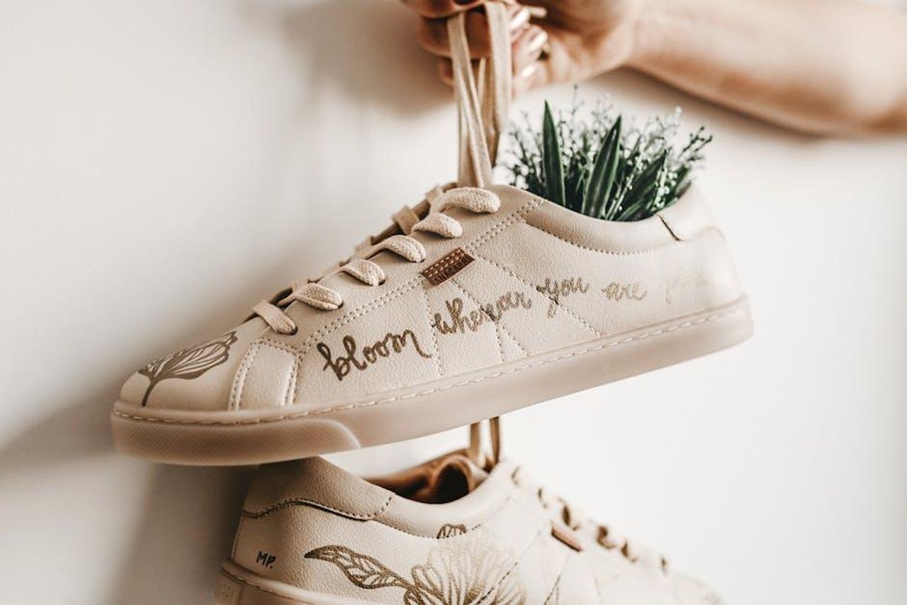 Sneakers_ispirate_ai_dolci_di_Milk_Bar_P8qSlATdpL - Confidenze24 Source: Jonathan Borba - pexels - Close-up of beige sneakers with plant design and 'bloom where you are planted' script.