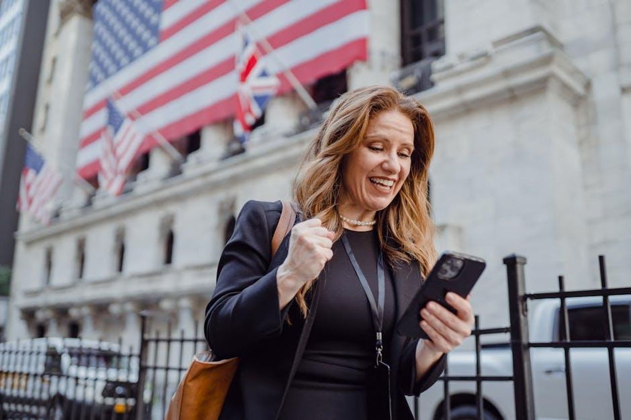 Toro_20_aprile__20_maggio_7hk7oGjROu - Confidenze24 Source: Mizuno K - pexels - Smiling woman celebrates success while checking mobile phone near iconic city building with flags.
