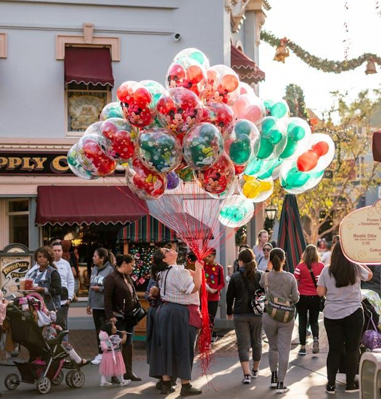 Tradizioni_familiari_a_Disneyland_zTRoX9nygi - Confidenze24 Source: Rafael Mendoza - pexels - A lively street scene featuring a vendor selling vibrant balloons amidst a bustling crowd during daytime festivities.