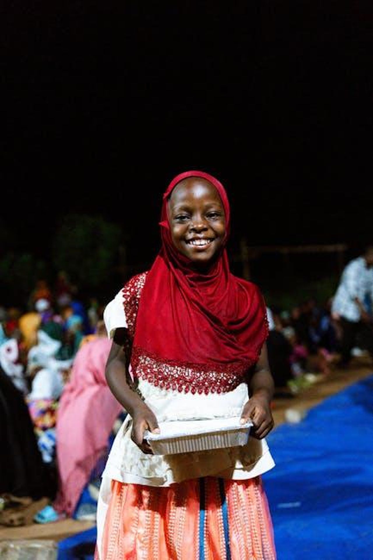 Trasformazione_della_charity_FnDrwYQeRb - Confidenze24 Source: Şeyhmus Kino - pexels - A joyful girl with red hijab holds a container at night outdoor event.