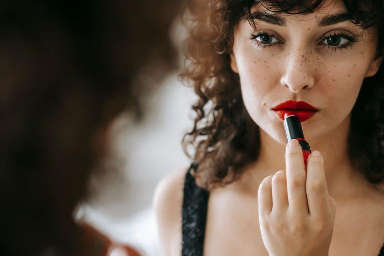 Un_alleato_per_il_trucco_quotidiano_R9XOQOLD3b - Confidenze24 Source: Sam Lion - pexels - Attractive female with freckles and dark curly hair looking at mirror while applying red lipstick during cosmetic routine in room