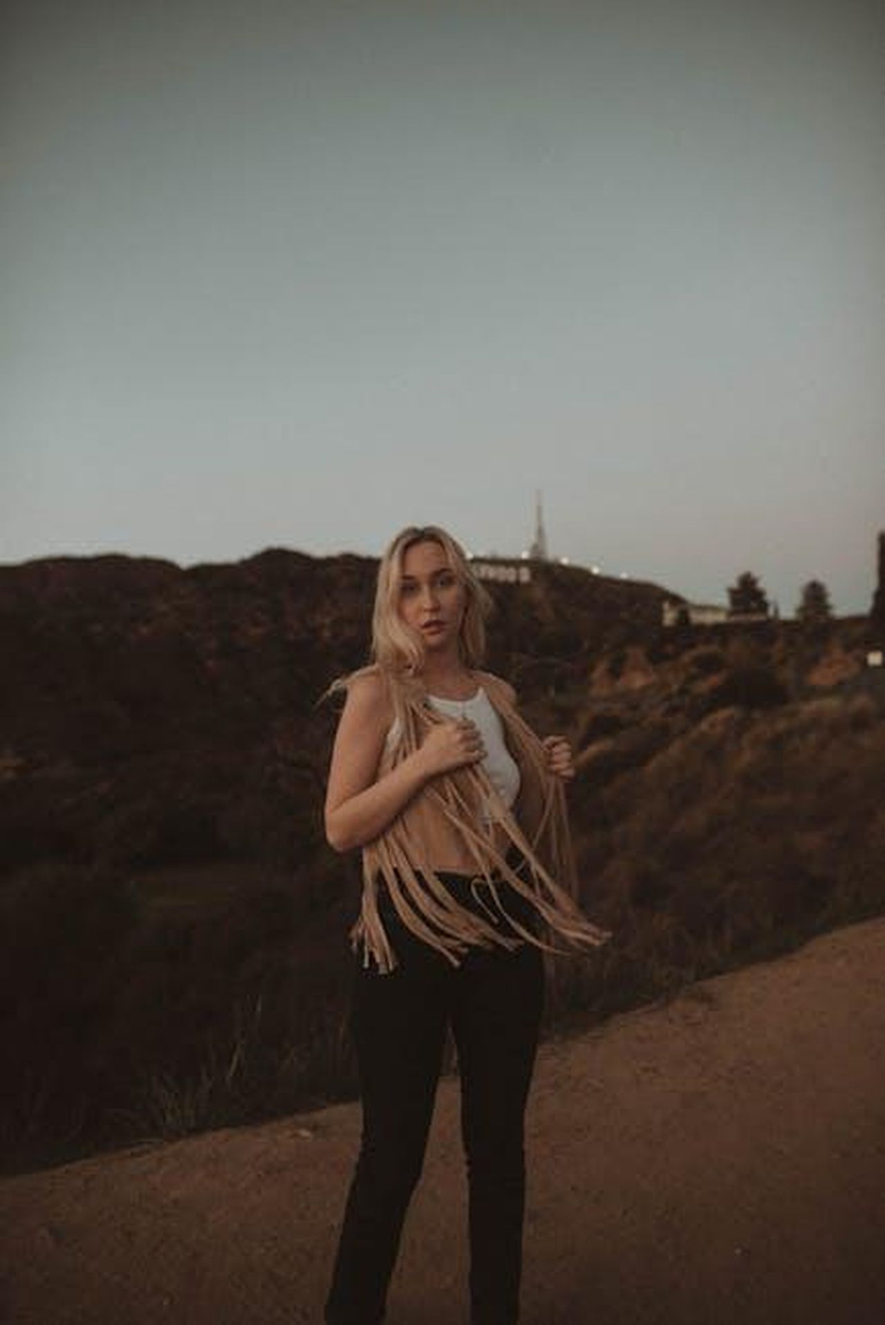 Una_notte_di_passione_con_la_star_di_Hollywood_Brigitte_Nielsen_BnIKXhL6mH - Confidenze24 Source: Nina Hill - pexels - A blonde woman in a fringe vest posing outdoors at twilight with a scenic landscape backdrop.