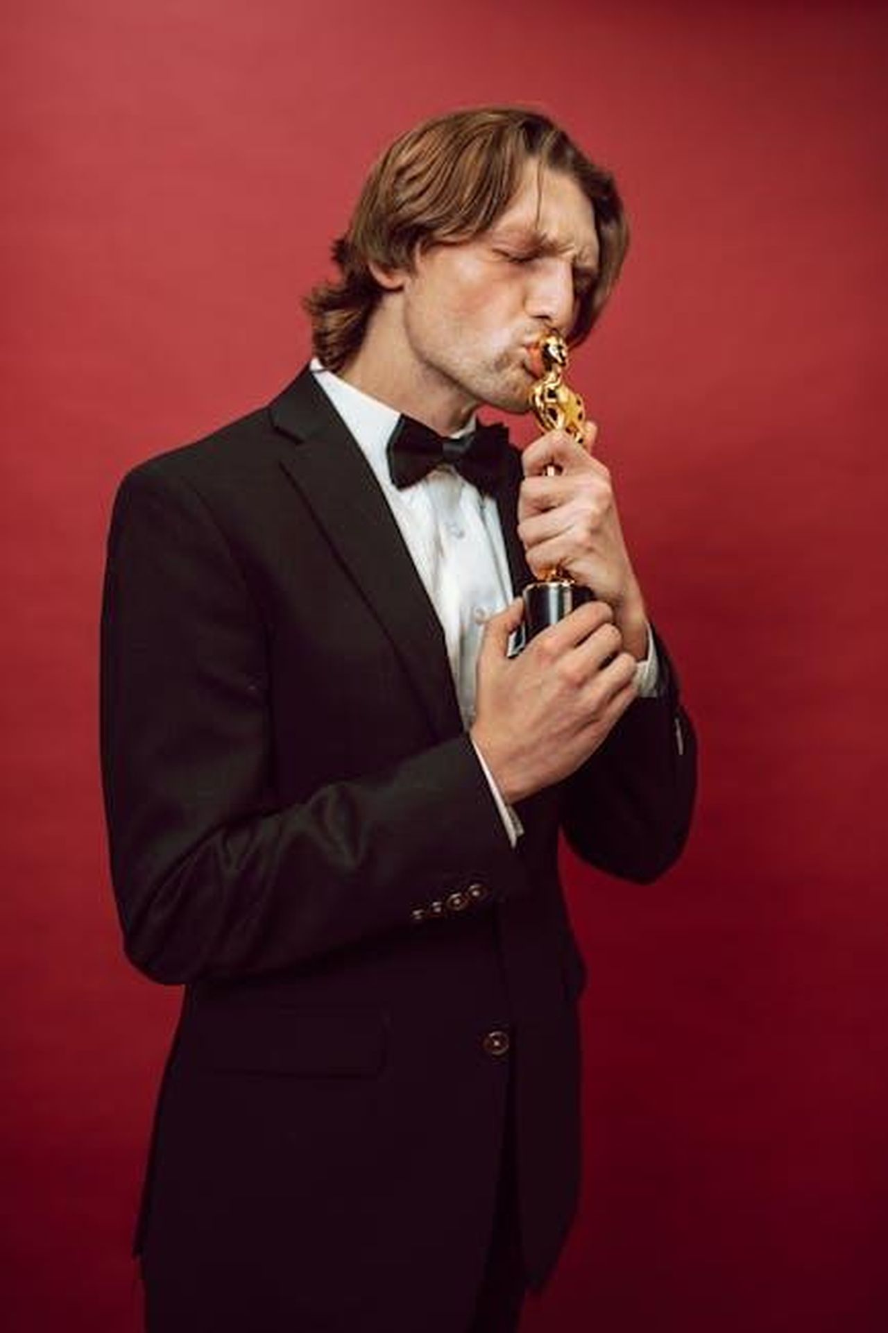 Vincitori_delle_categorie_principali_eA3kbWX02o - Confidenze24 Source: RDNE Stock project - pexels - Man in a tuxedo kissing a trophy, symbolizing achievement, against a vibrant red background.