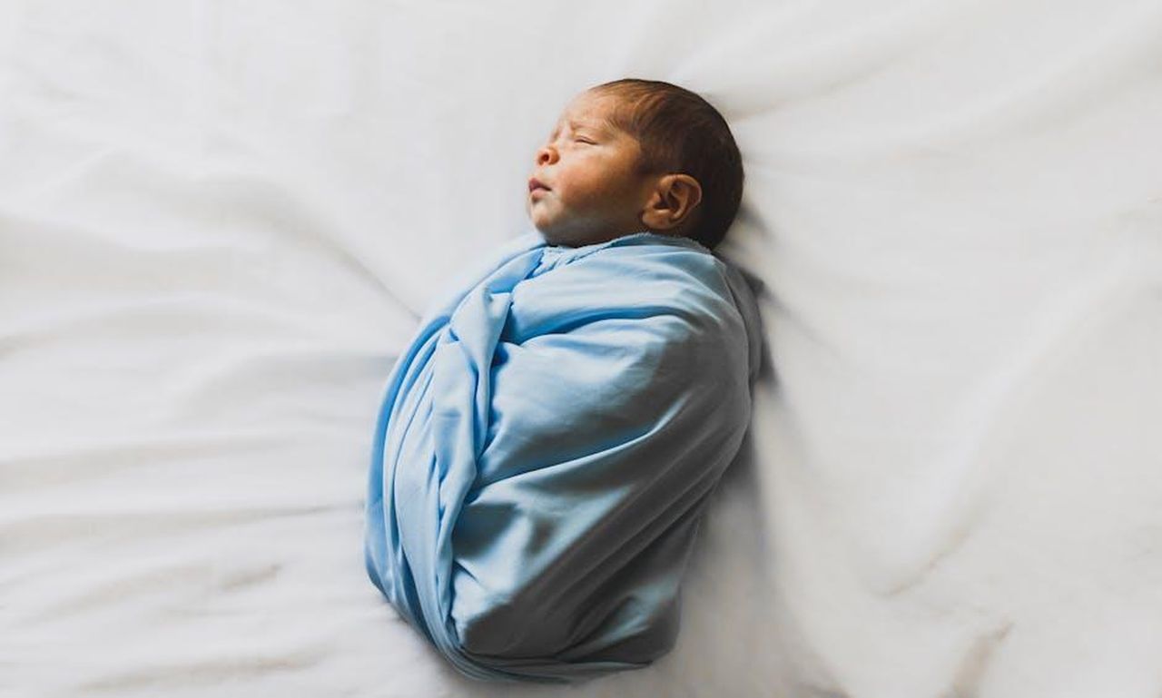 Source: Laura Garcia - pexels - A serene newborn baby wrapped in a blue blanket, sleeping peacefully indoors.