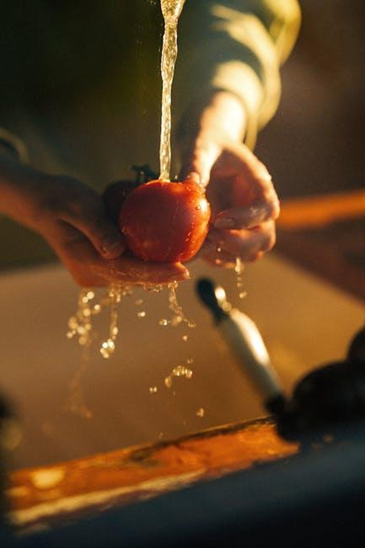 Tecniche_per_far_pulire_i_bambini_BQZ5LYVHgD - Confidenze24 Source: Gül Işık - pexels - A person washing a fresh tomato under a faucet in warm kitchen lighting, highlighting cleanliness and health.